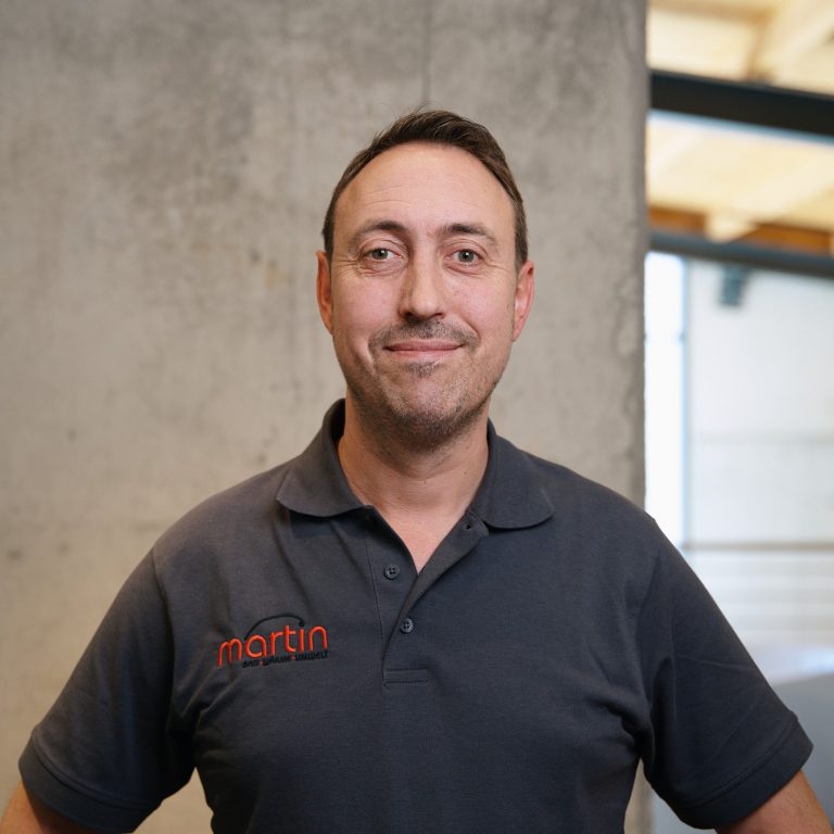 Bodo Neuhaus - Badgestalter Ein Mann steht vor einer Betonwand, trägt ein graues Shirt mit Logo und lächelt freundlich.