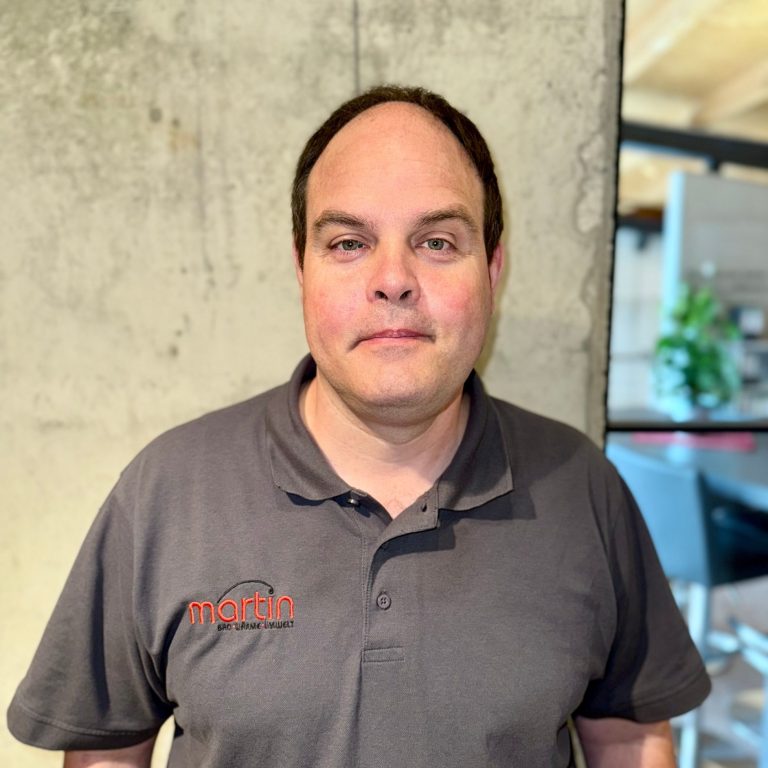 Bodo Neuhaus - Badgestalter Ein Mann steht vor einer Betonwand, trägt ein graues Shirt mit Logo und lächelt freundlich.